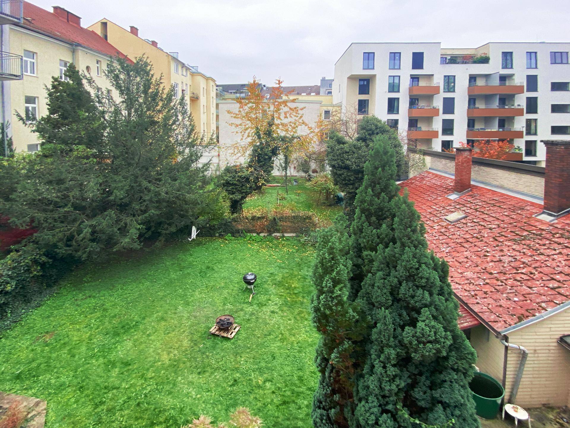 Grüner Gartenbereich mit Rasen, Bäumen und Grill, umgeben von Wohngebäuden.