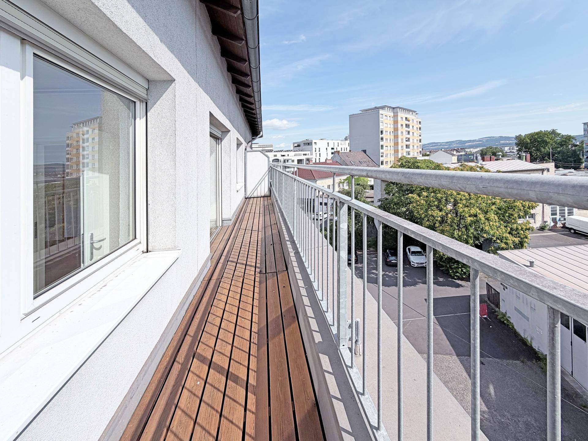 Langer Balkon mit Holzfußboden und weißem Geländer, bietet Blick auf die Straße und Gebäude.