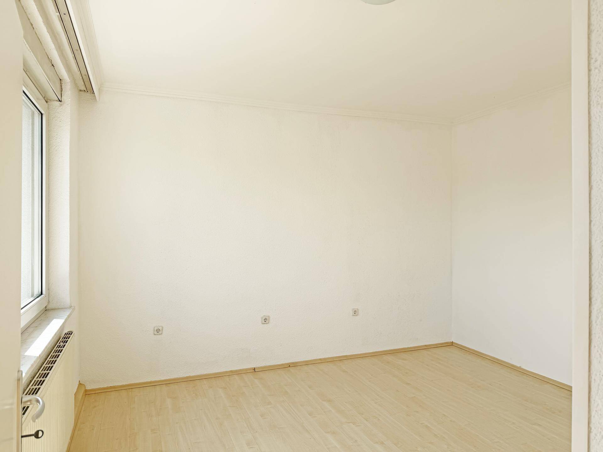 Leeres, helles Zimmer mit weißen Wänden, hellem Holzboden und einem Fenster.