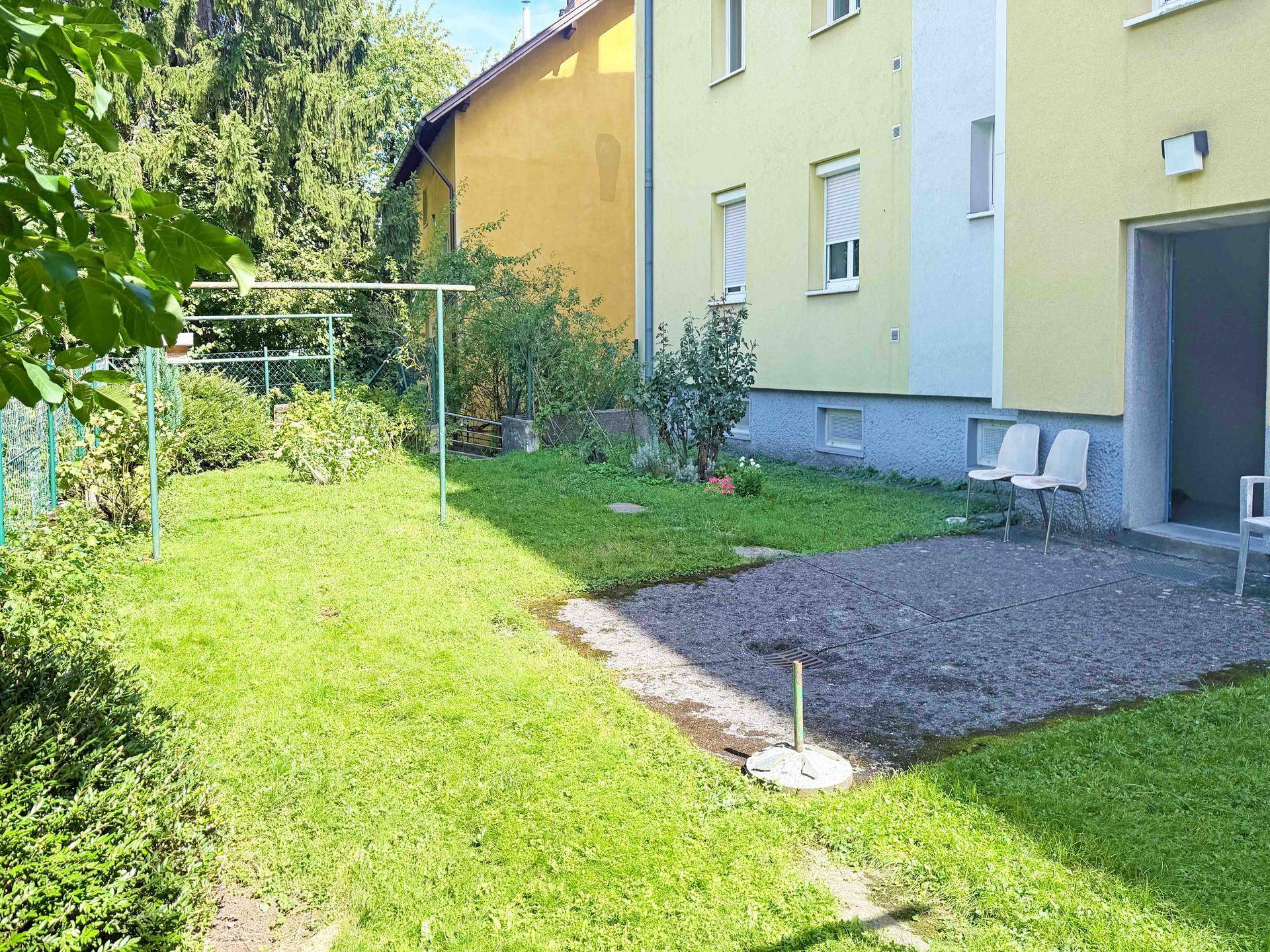 Grüner Gartenbereich mit Rasenfläche und kleiner Terrasse hinter dem Gebäude.