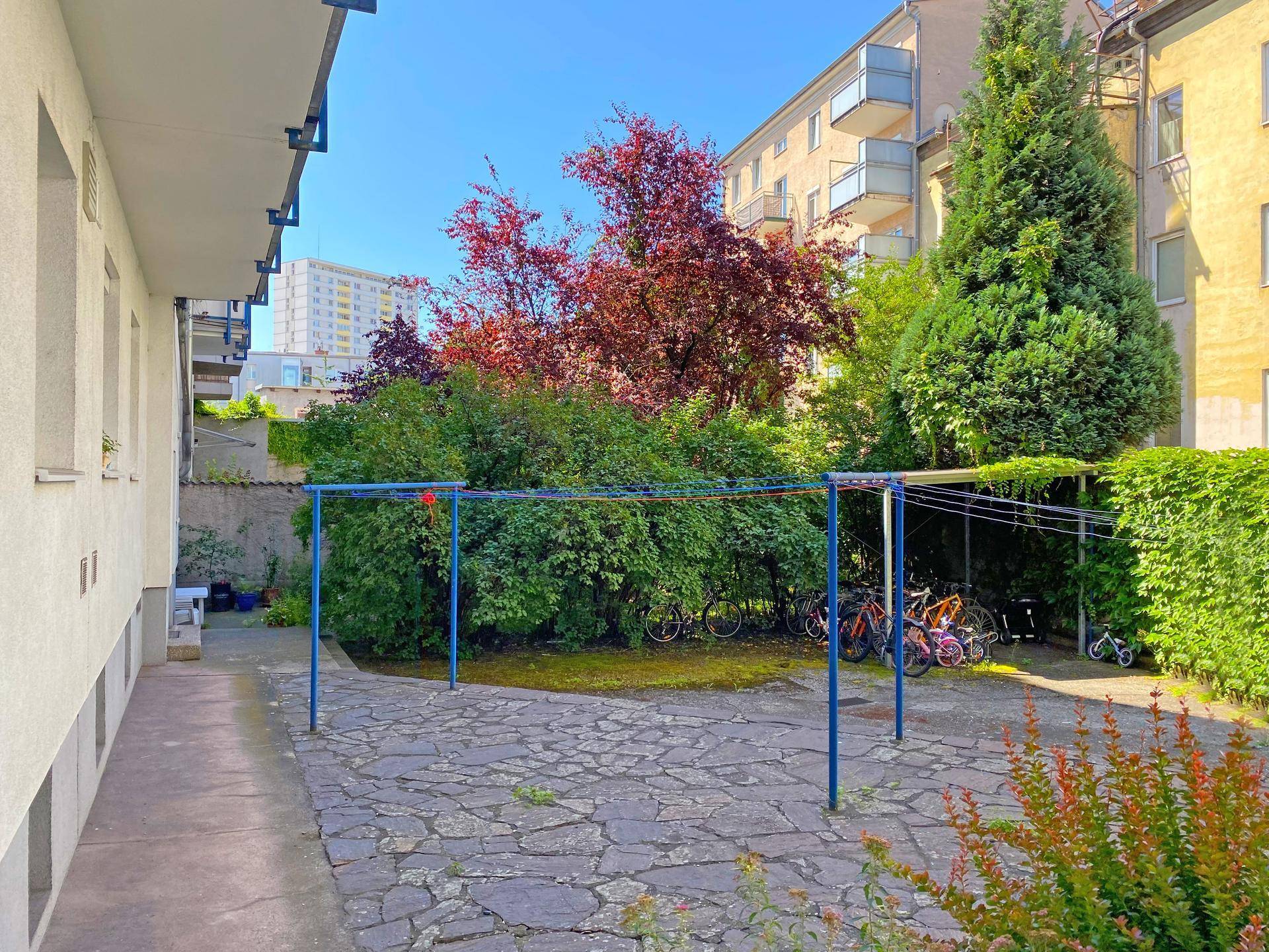 Außenbereich mit gepflasterter Terrasse, Rasenfläche, Bäumen und Fahrradabstellmöglichkeiten.