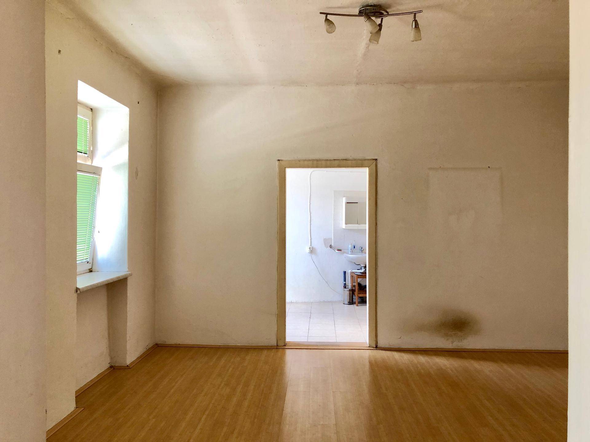 Unrenovierter Raum mit Holzboden und Blick durch eine offene Tür in ein Badezimmer.
