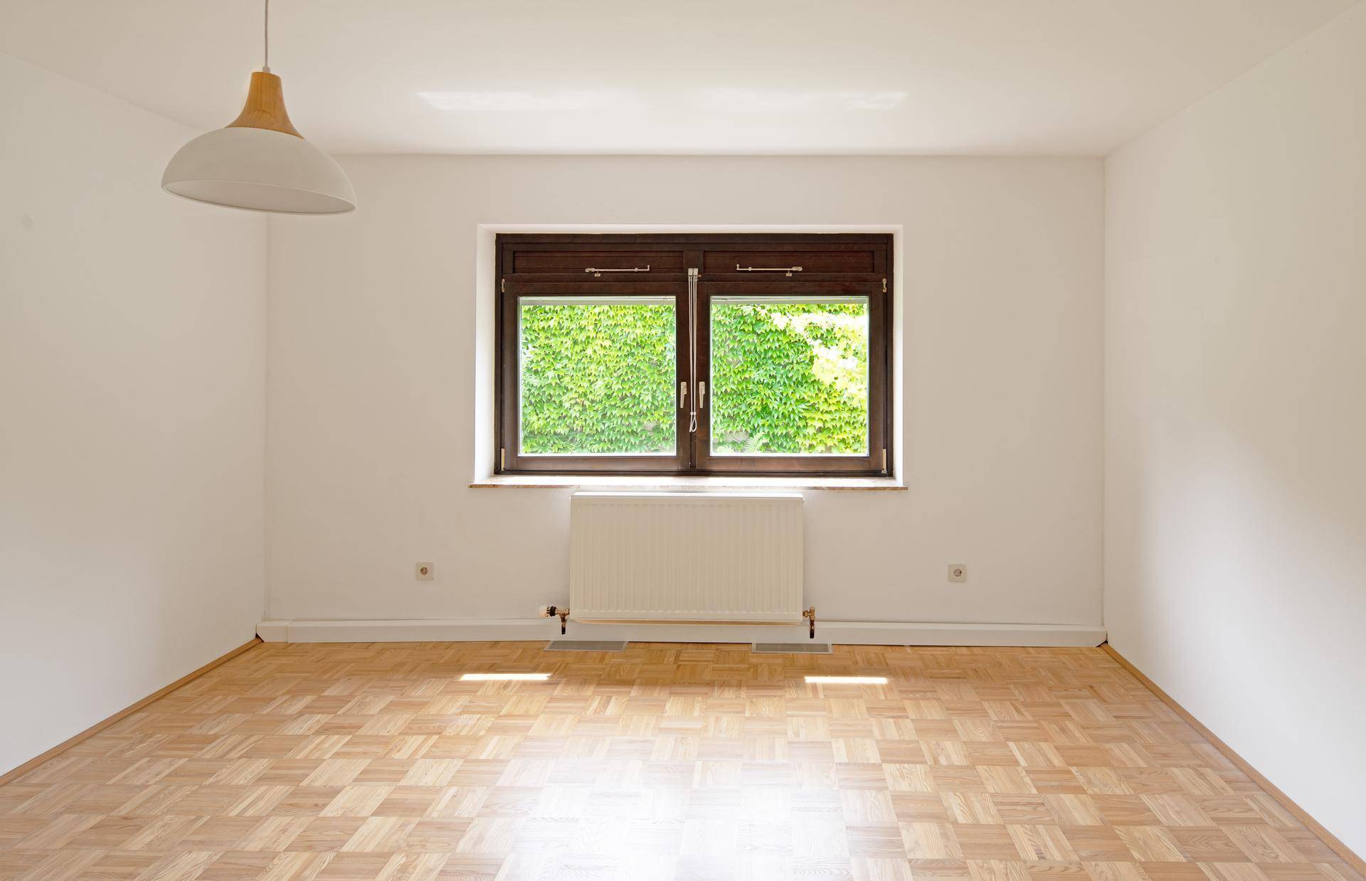 Freundliches Zimmer mit Parkettboden und zwei Fenstern, die viel Tageslicht hereinlassen und eine grüne Aussicht bieten.