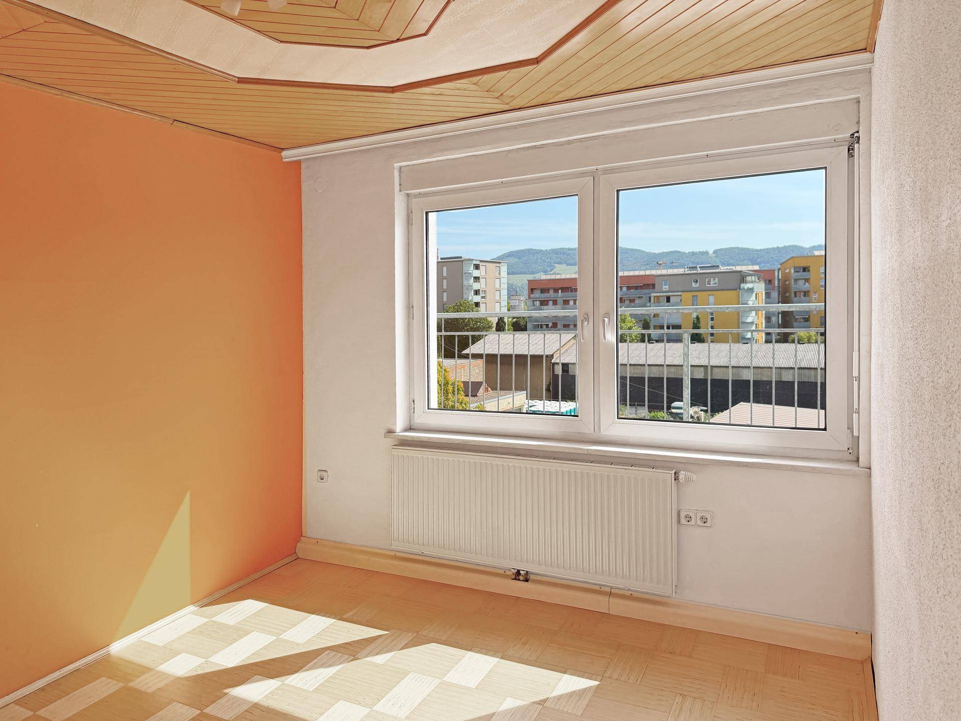 Helles Zimmer mit orangefarbenen Wänden, Holzdecke und Fenster mit Blick auf die Umgebung.