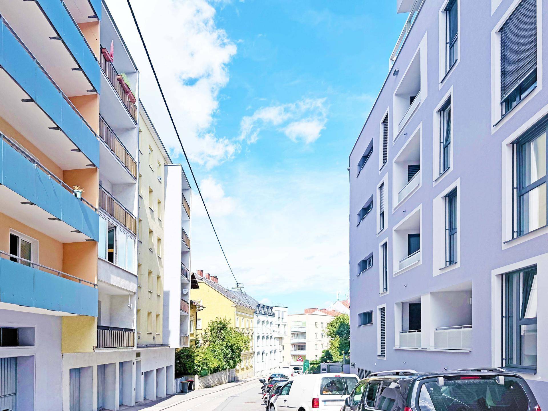 Straßenansicht mit verschiedenen Wohngebäuden und geparkten Autos in urbaner Umgebung.