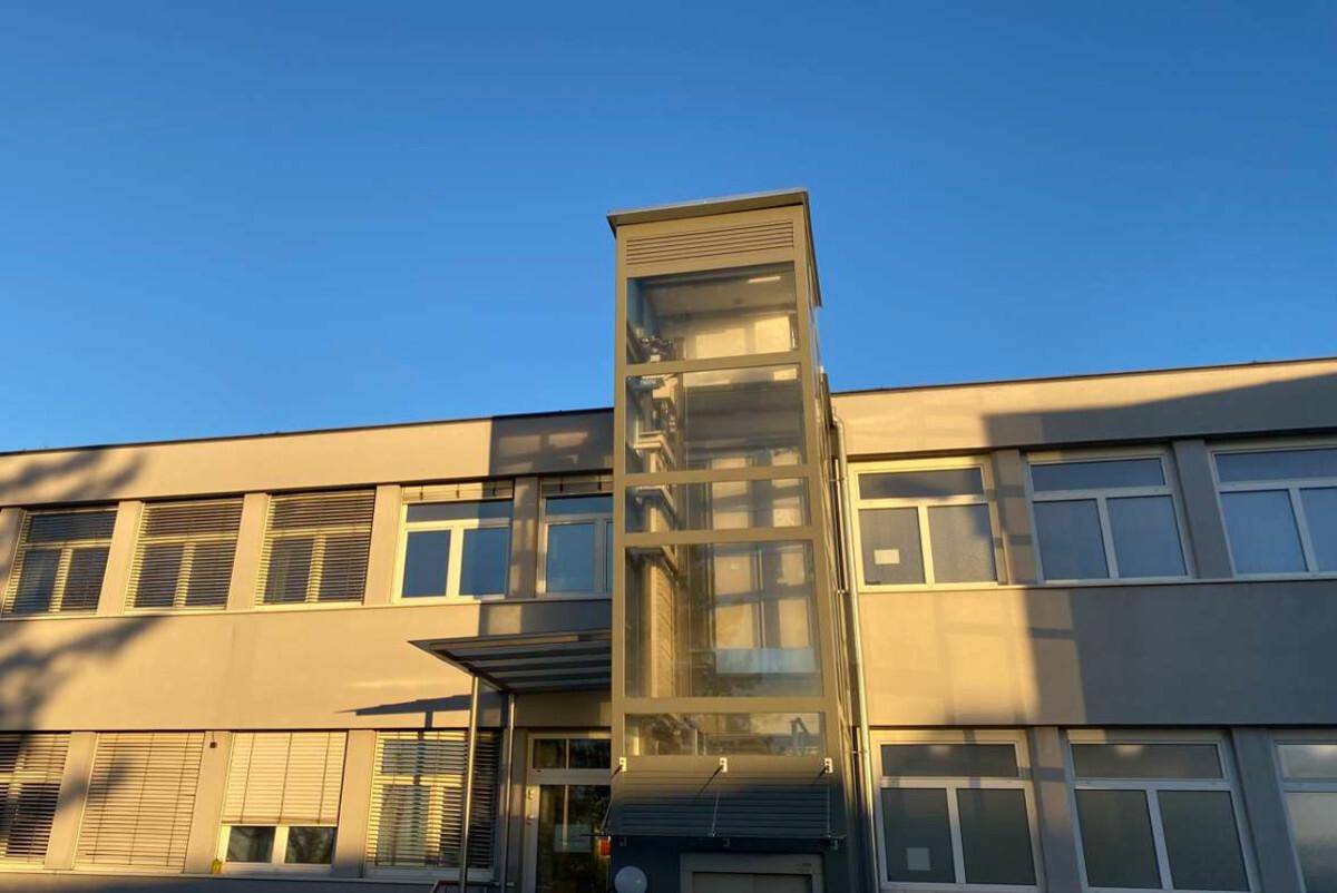 Nahaufnahme des gläsernen Aufzugs an der Fassade des Bürogebäudes bei Sonnenuntergang mit klarem Himmel.