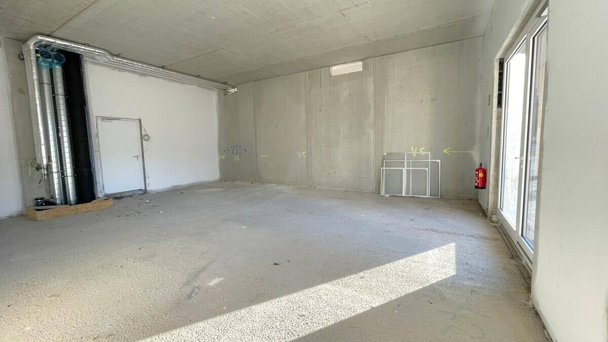 Geräumiger Gewerberaum im Rohbau mit Betonboden, großen Fenstern und sichtbaren Versorgungsleitungen.