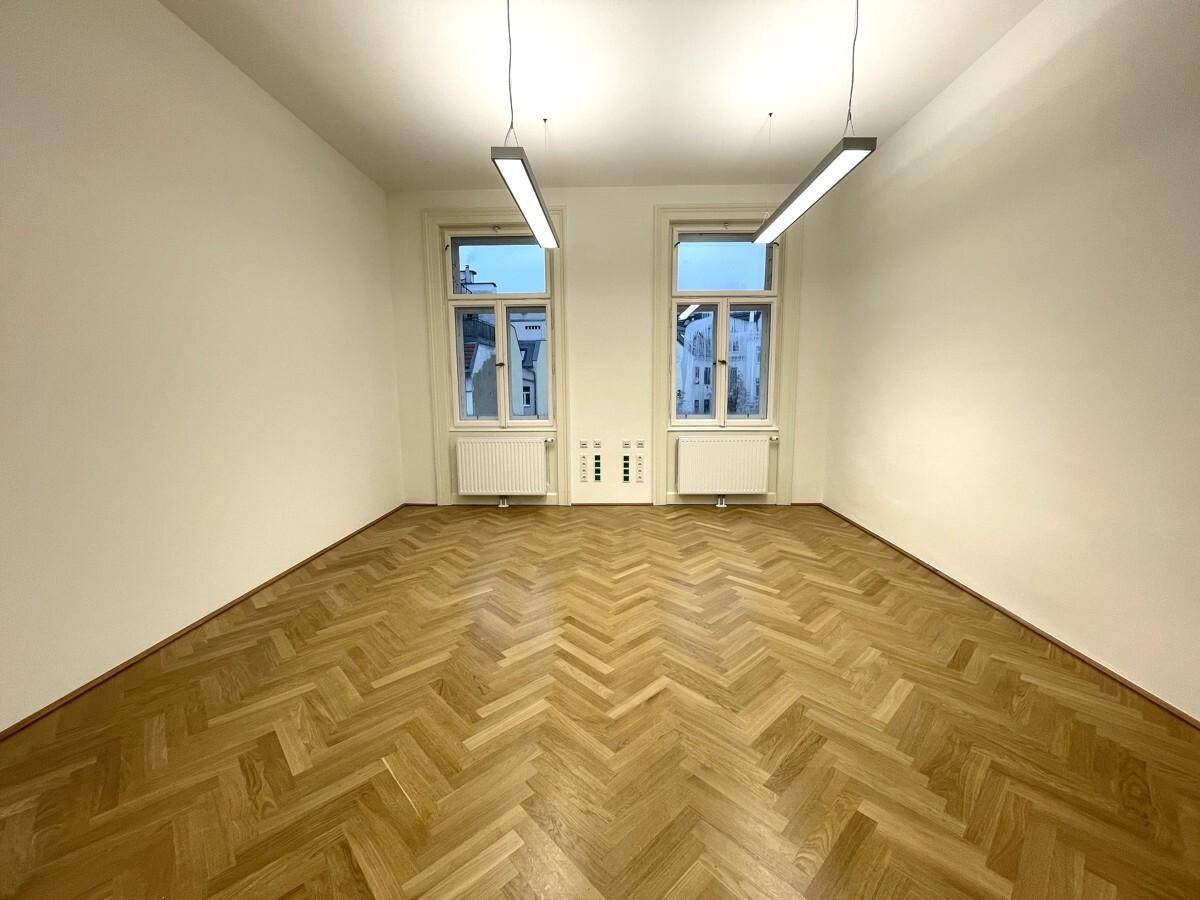 Heller Büroraum mit Fischgrätparkett und zwei großen Fenstern, die eine offene und einladende Atmosphäre schaffen.