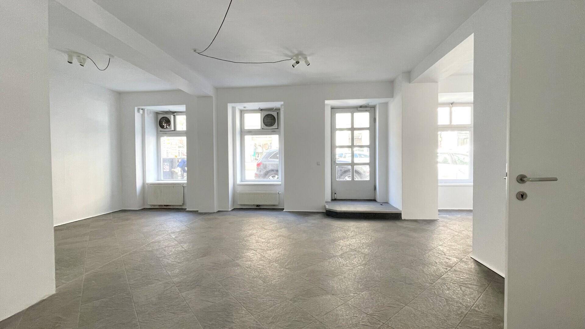 Heller, leerer Raum mit mehreren großen Fenstern zur Straße und grauen Fliesen, bietet viel natürliches Licht.
