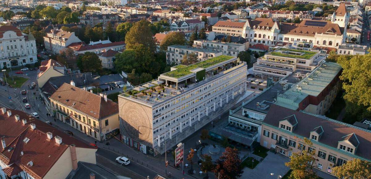 Luftaufnahme eines modernen Gebäudes mit begrüntem Dach inmitten einer städtischen Umgebung und umliegenden Häusern.