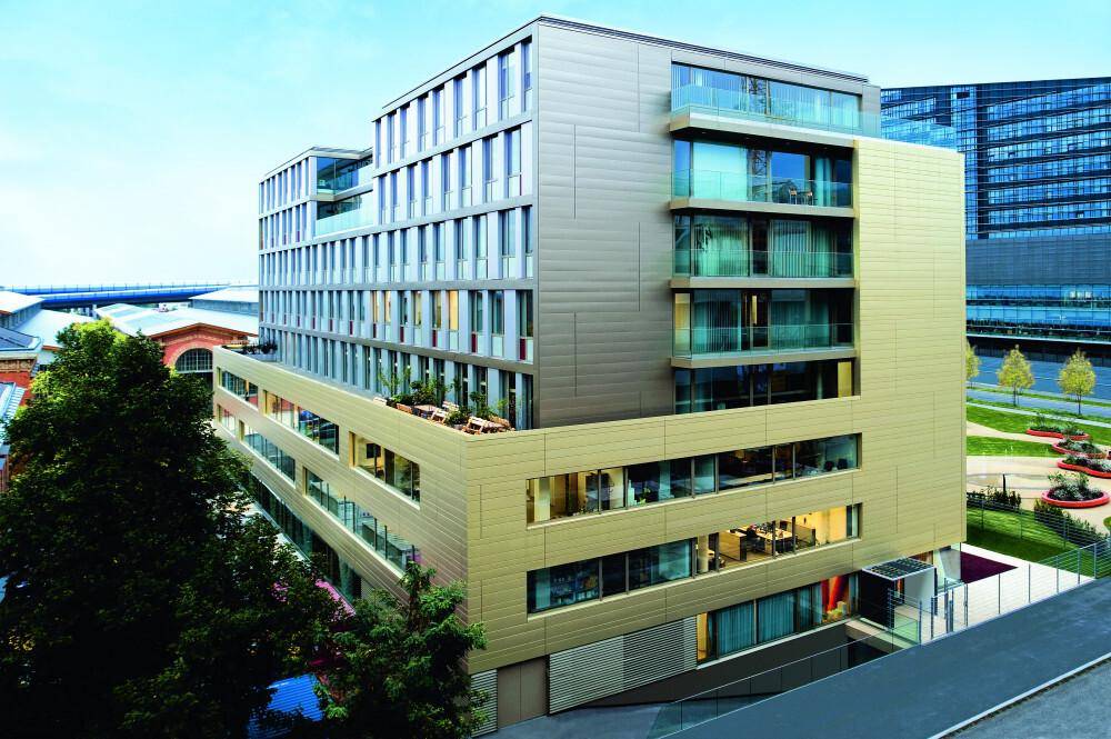 Modernes Bürogebäude mit mehreren Etagen, großen Fenstern und Balkonen in städtischer Umgebung, unter blauem Himmel.