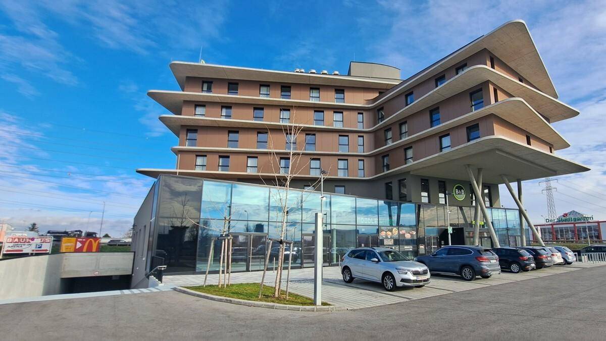 Modernes Gebäude mit geschwungenen Etagen, Glasfassade und Zufahrt zu einer Tiefgarage, umgeben von Parkplätzen.