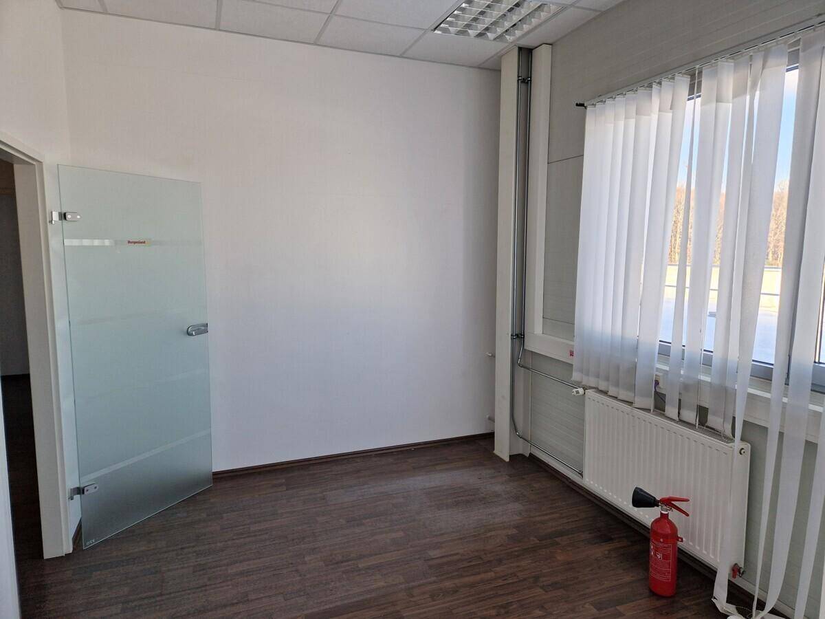 Leerer Raum mit dunklem Laminatboden, weißen Wänden, einem Fenster mit Jalousien und einem Heizkörper.