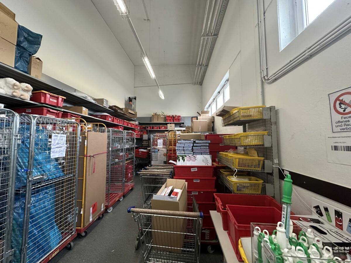 Lagerraum mit hohen Regalen, Rollcontainern und Kartons, beleuchtet durch Deckenleuchten und kleine Fenster.