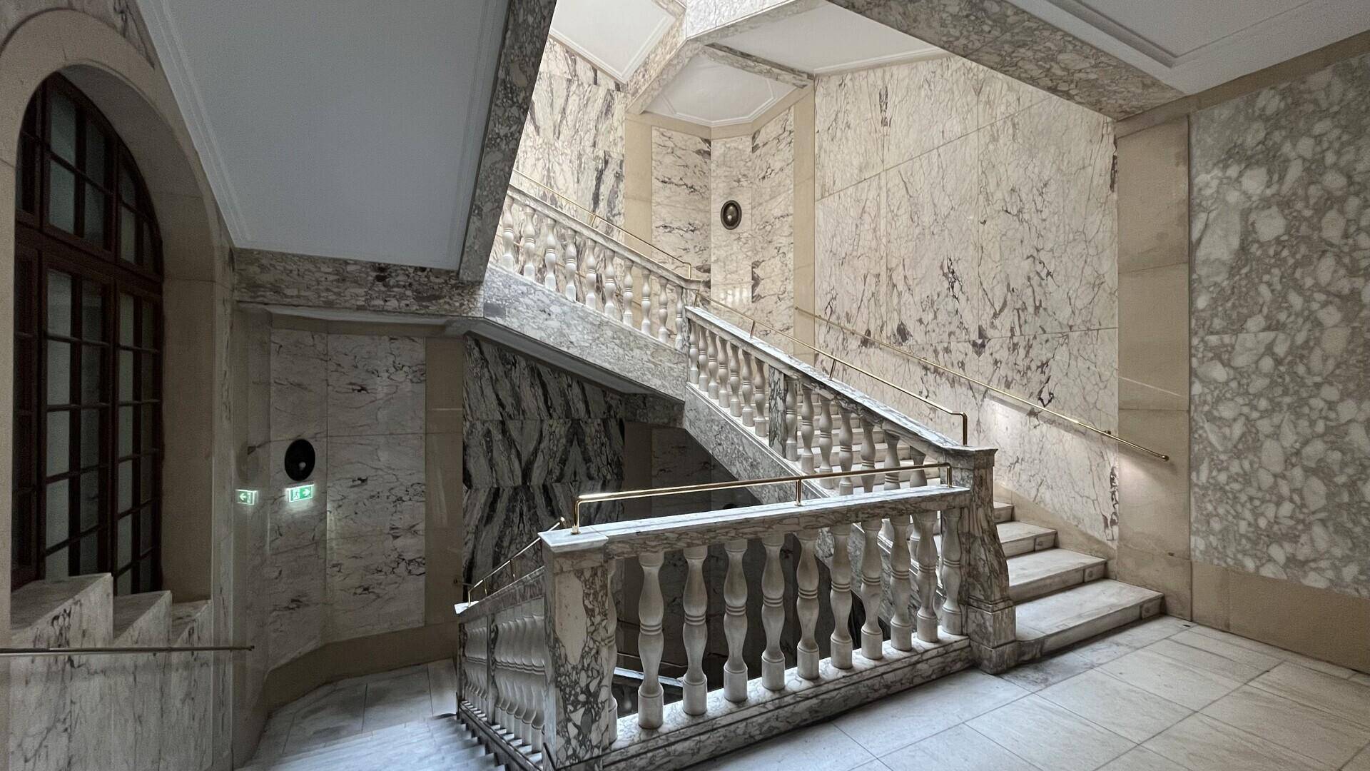Prunkvolles Treppenhaus mit Marmorstufen und -geländer, hohen Wänden und einem großen Bogenfenster.
