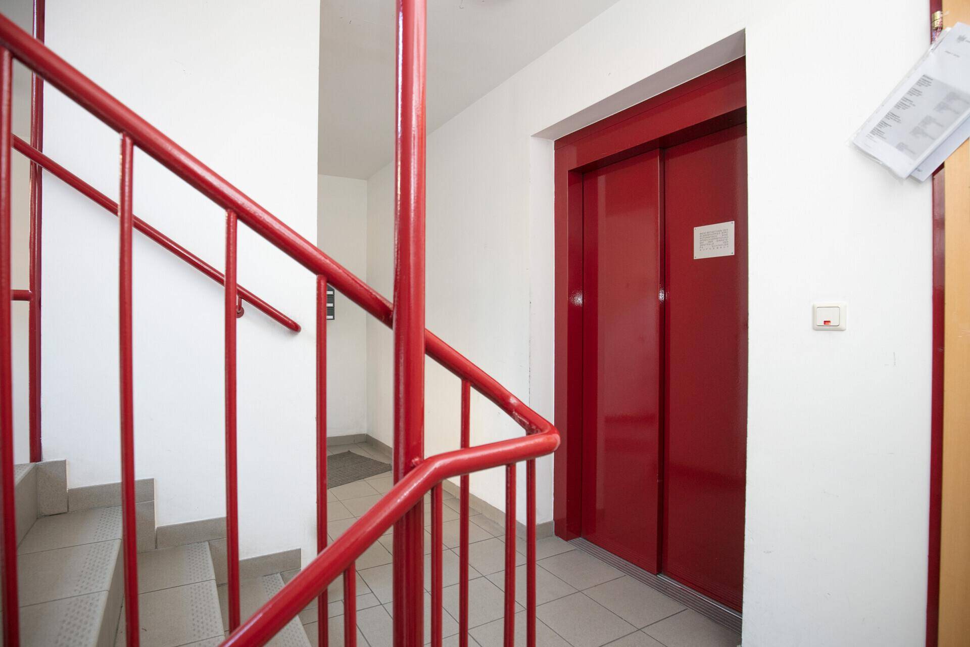 Treppenhaus mit rotem Geländer und einem modernen roten Aufzug, der alle Etagen verbindet.