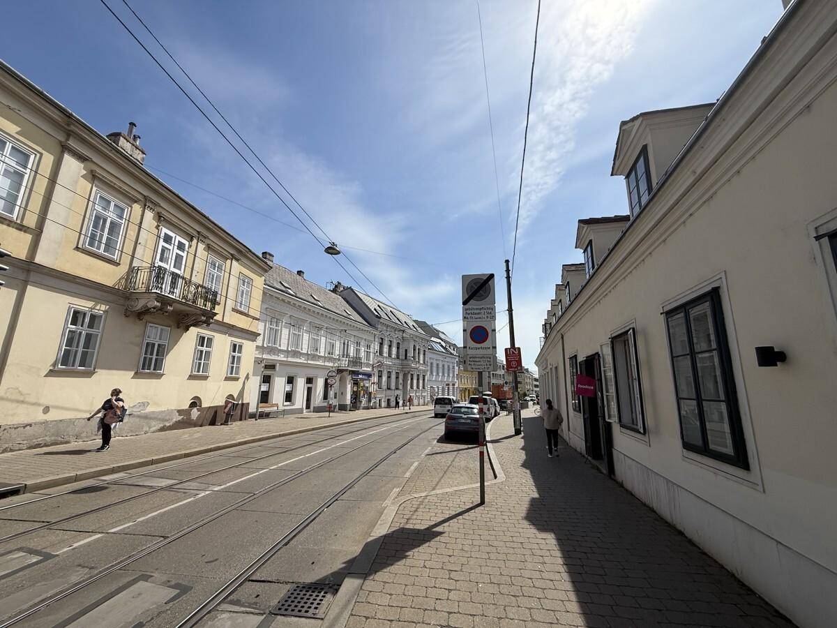 Eine belebte Straße mit historischen Gebäuden, Straßenbahnschienen und Fußgängern unter blauem Himmel.