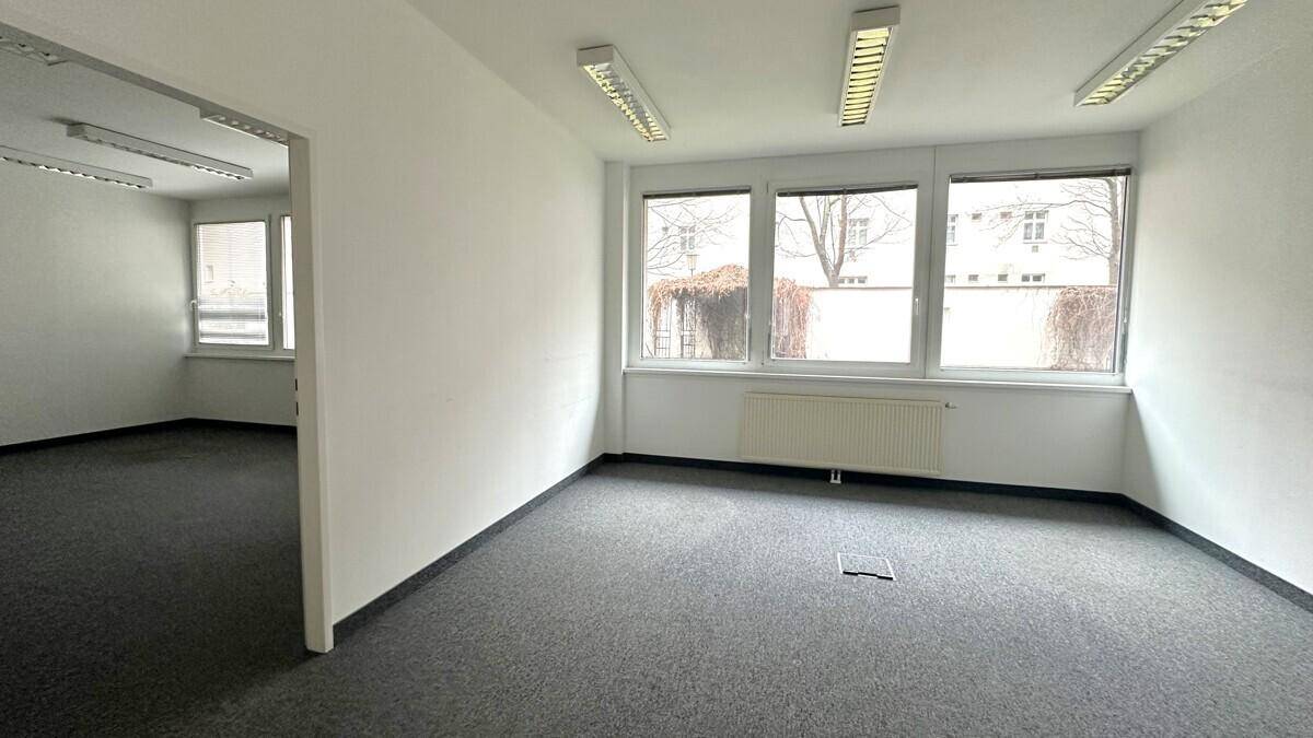 Offener Bürobereich mit grauem Teppichboden, weißen Wänden und großen Fenstern, die viel Licht spenden.
