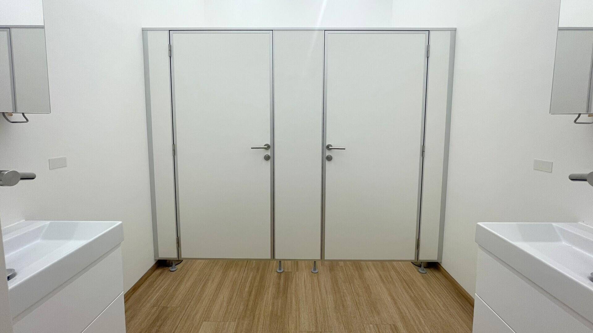 Moderner Toilettenbereich mit zwei Kabinen, Waschbecken und Holzboden in hellem, funktionalem Design.