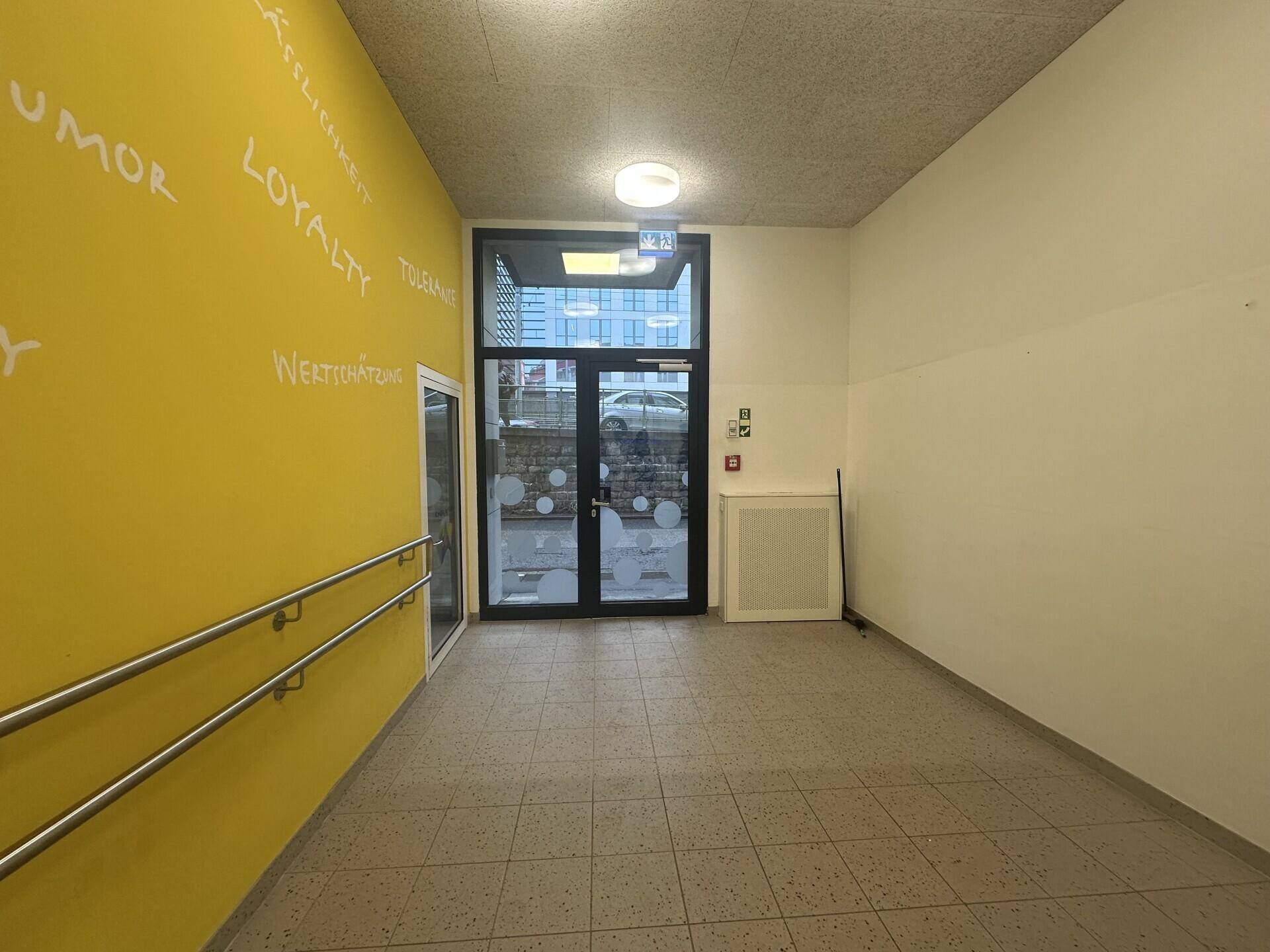 Heller Eingangsbereich mit gelber Wand, Fliesenboden und Glastüren, die nach außen führen.