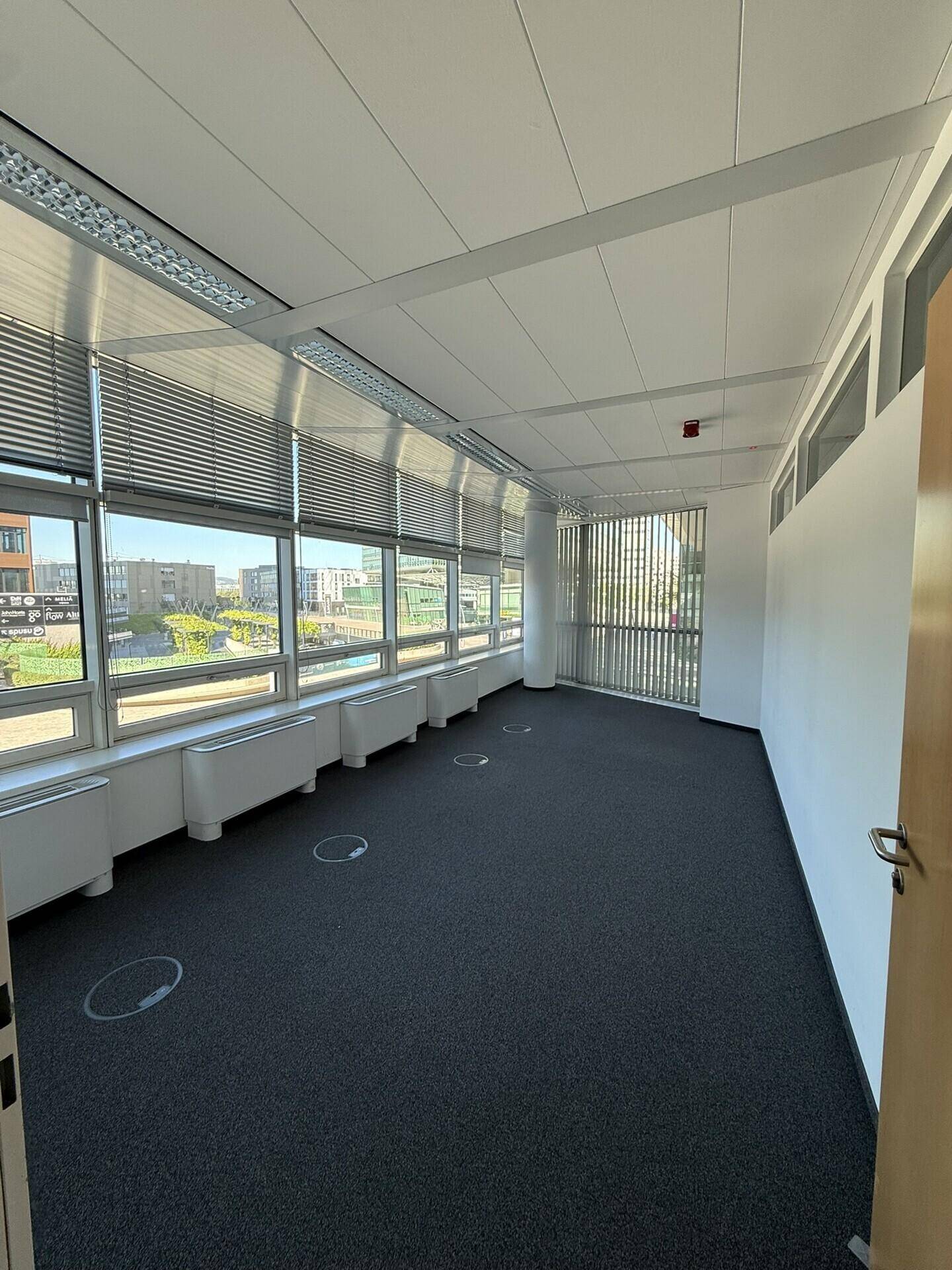 Helles Büro mit Teppichboden, breiter Fensterfront und Jalousien, die den Blick nach draußen freigeben.