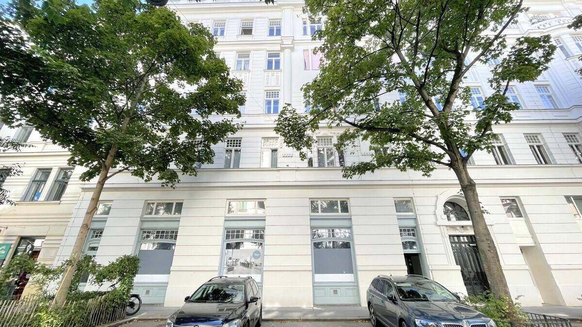 Fassade eines historischen Gebäudes mit großen Fenstern im Erdgeschoss und parkenden Autos.