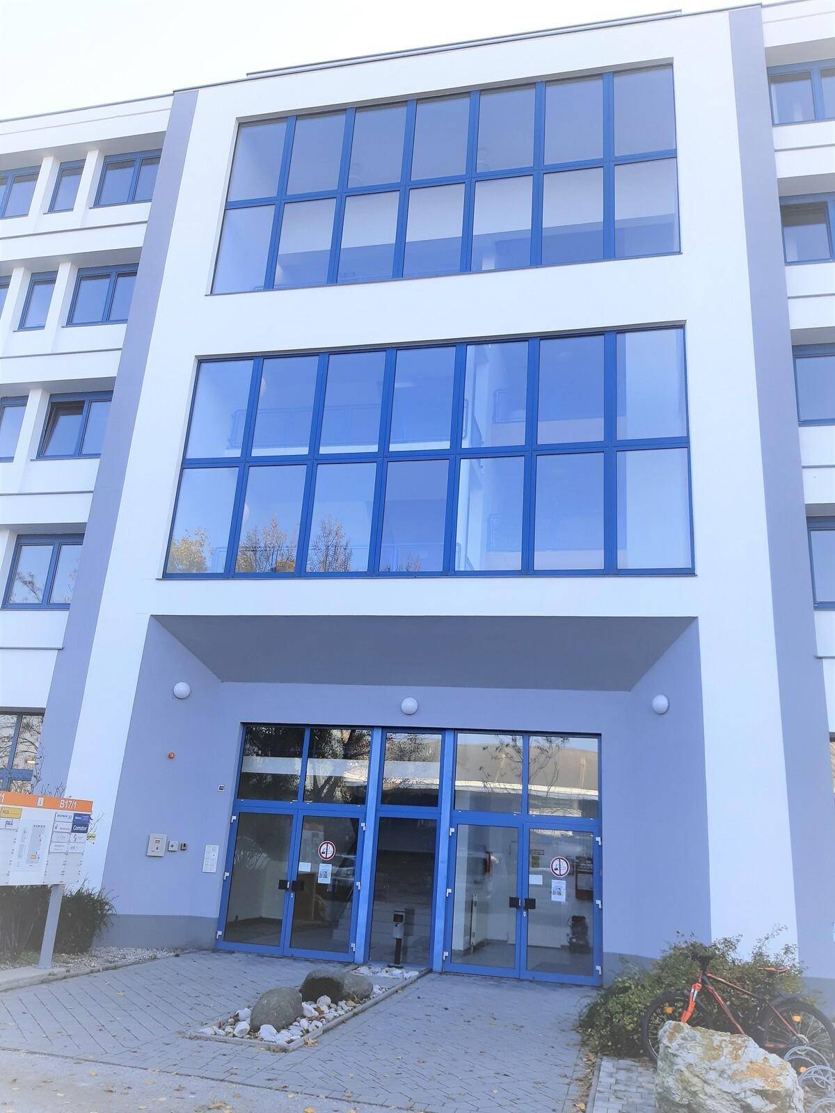 Haupteingang eines Bürogebäudes mit großen Glasfronten und blauen Türrahmen, umgeben von heller Fassade.