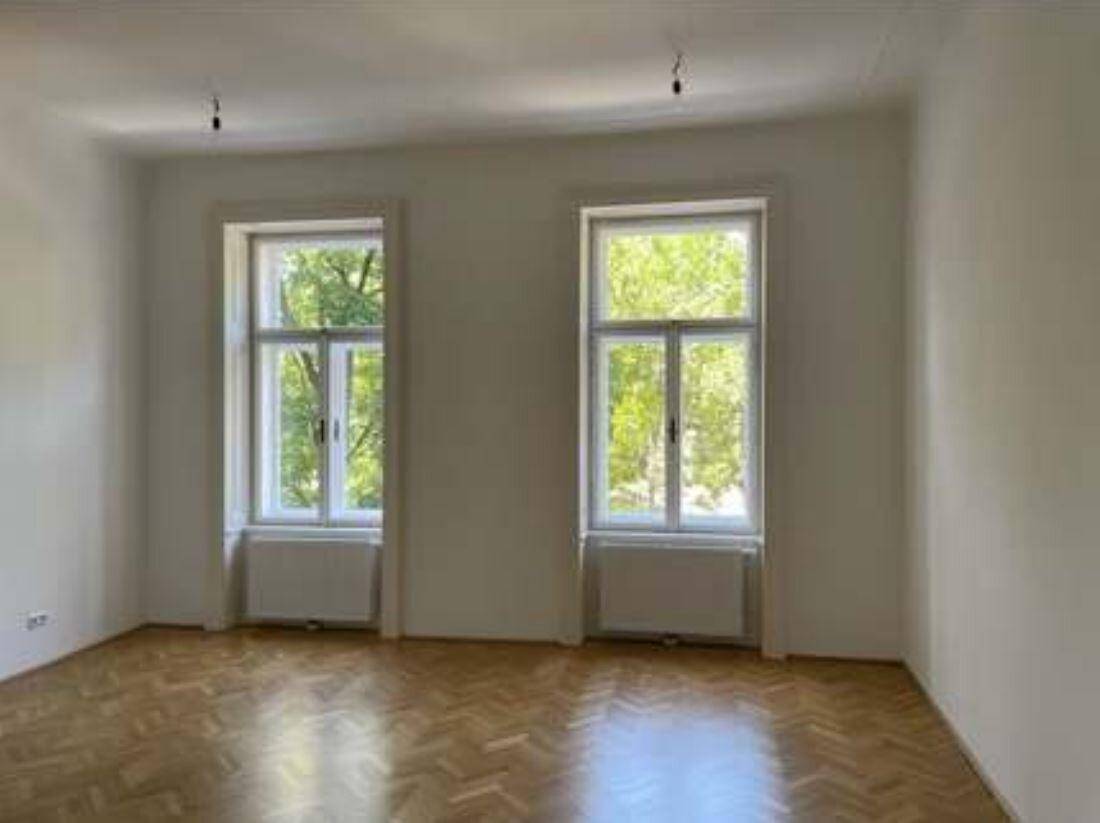 Leerer Raum mit Fischgrätparkett und zwei hohen Fenstern, die viel Tageslicht hereinlassen.