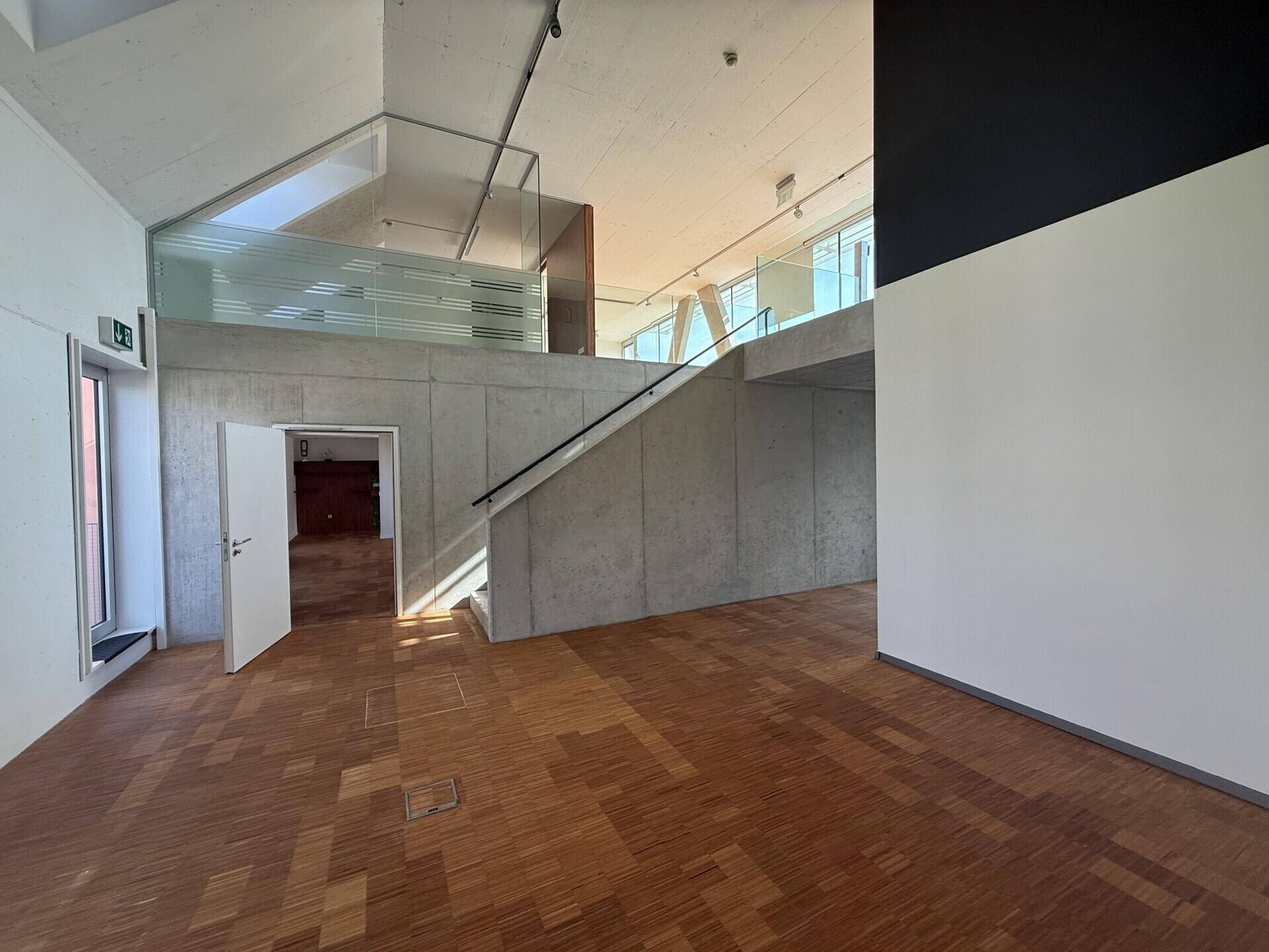 Geräumiger Bürobereich mit Holzboden, Sichtbetondecke und einer modernen Treppe mit Glasgeländer.