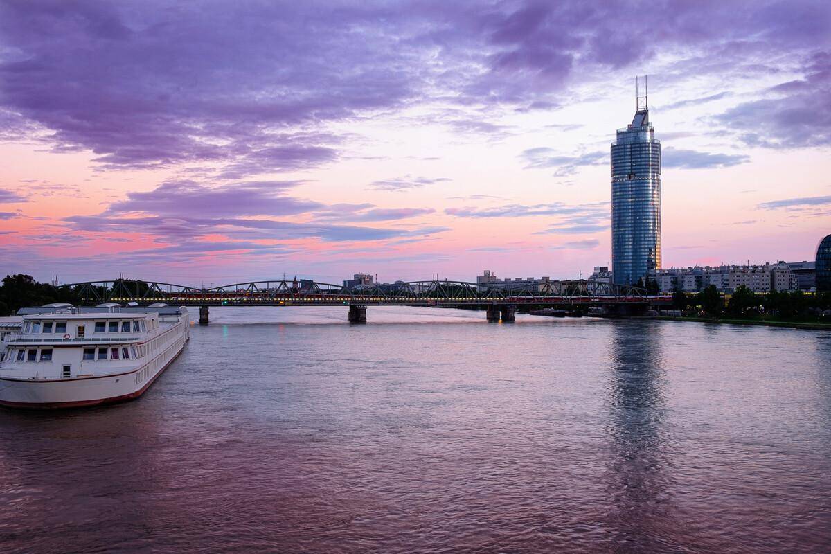 Abendliche Ansicht der Donau mit einem Kreuzfahrtschiff, beleuchteter Brücke und einem Hochhaus unter violettem Himmel.