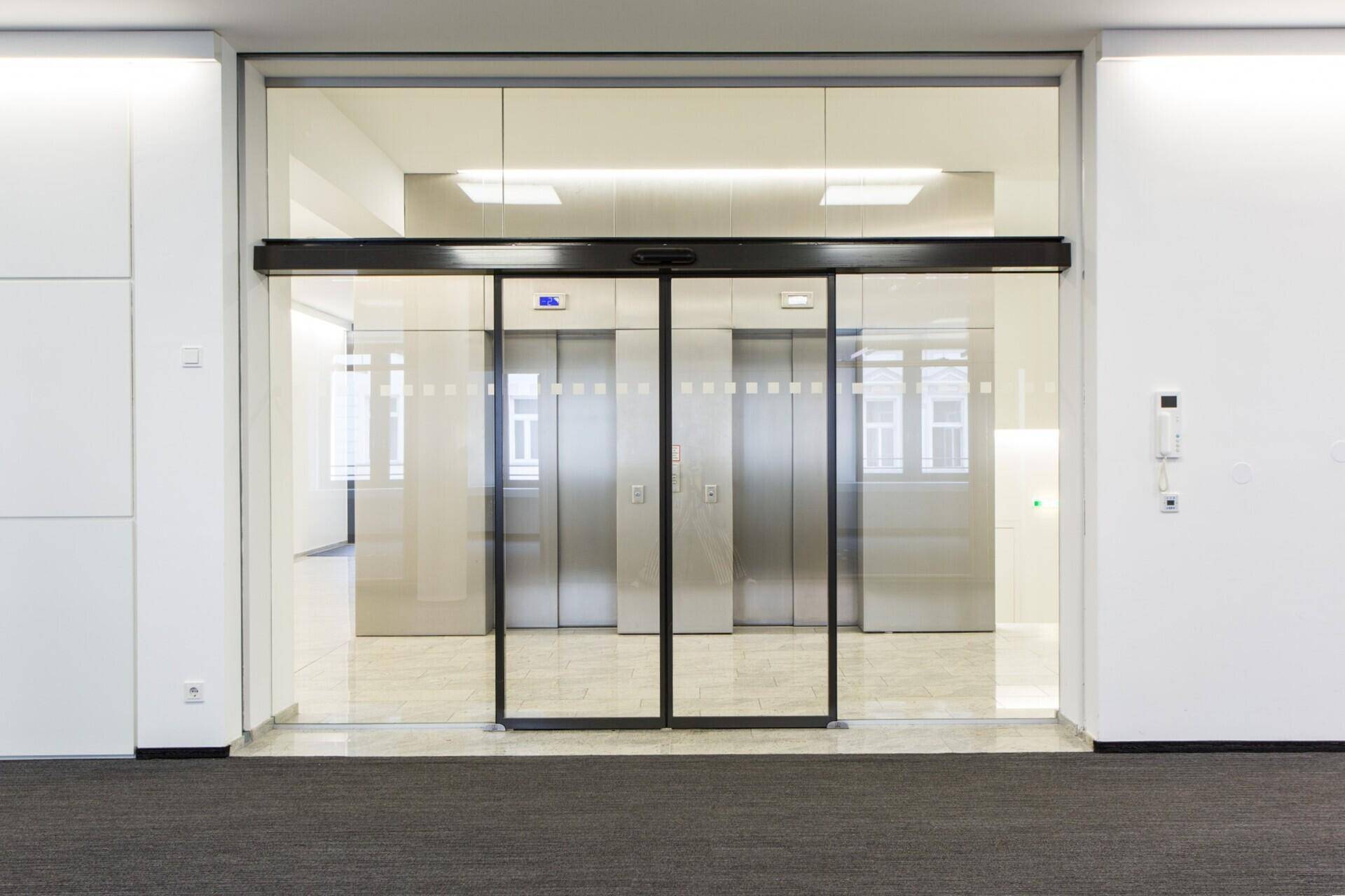 Eingangsbereich mit automatischen Glastüren und zwei modernen Aufzügen, die Zugang zu den Etagen bieten.