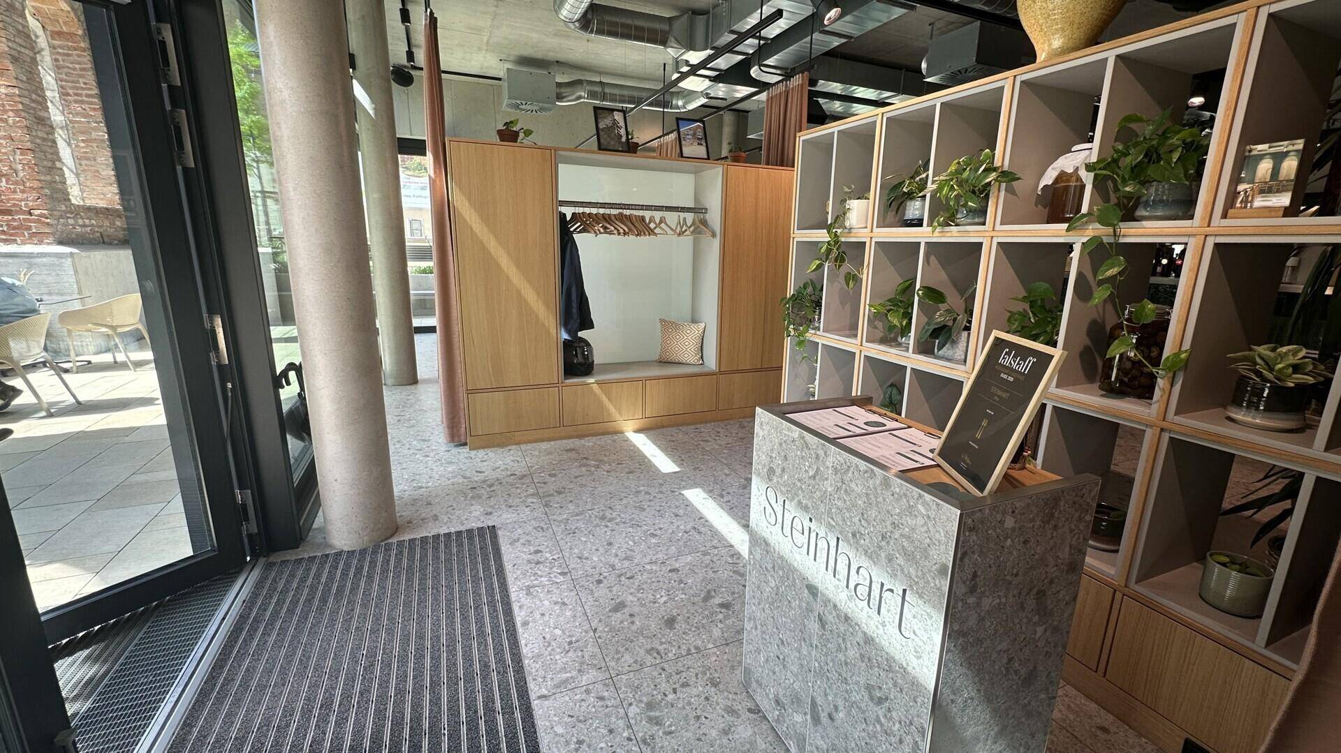 Moderner Eingangsbereich mit Empfangstheke, Garderobe und Regalen voller Pflanzen, auf Terrazzoboden.