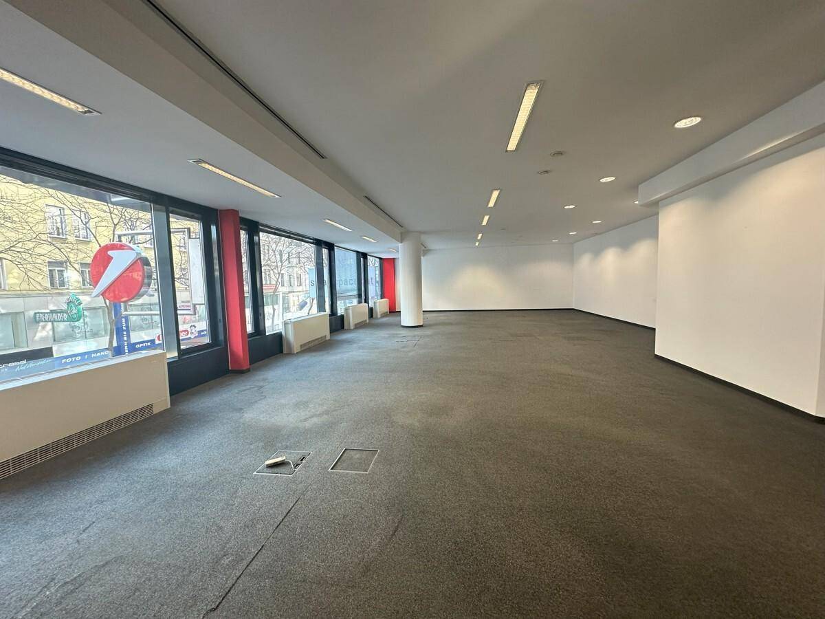 Geräumiger, leerer Bürobereich mit durchgehenden Fenstern und grauem Teppichboden, ideal für flexible Gestaltung.