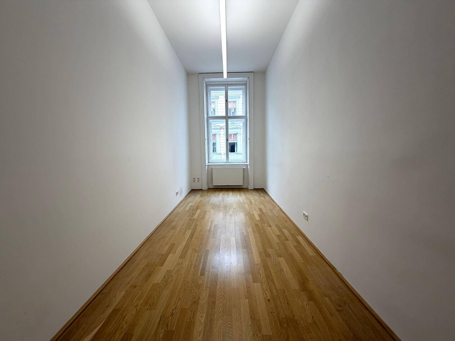 Langer, schmaler Raum mit Holzboden, einem Fenster am Ende und weiß gestrichenen Wänden.