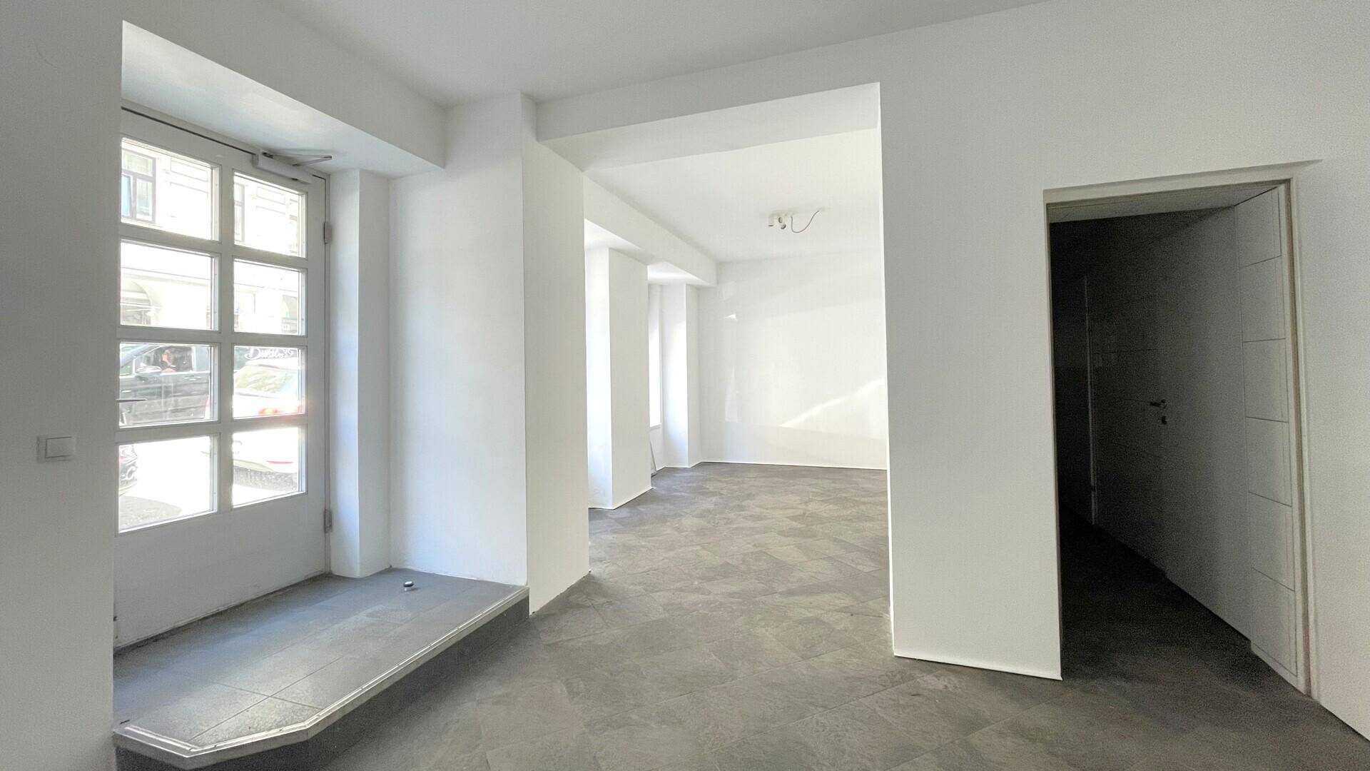Eingangsbereich mit einer großen Glastür zur Straße und grauen Fliesen, führt in einen hellen, offenen Raum.
