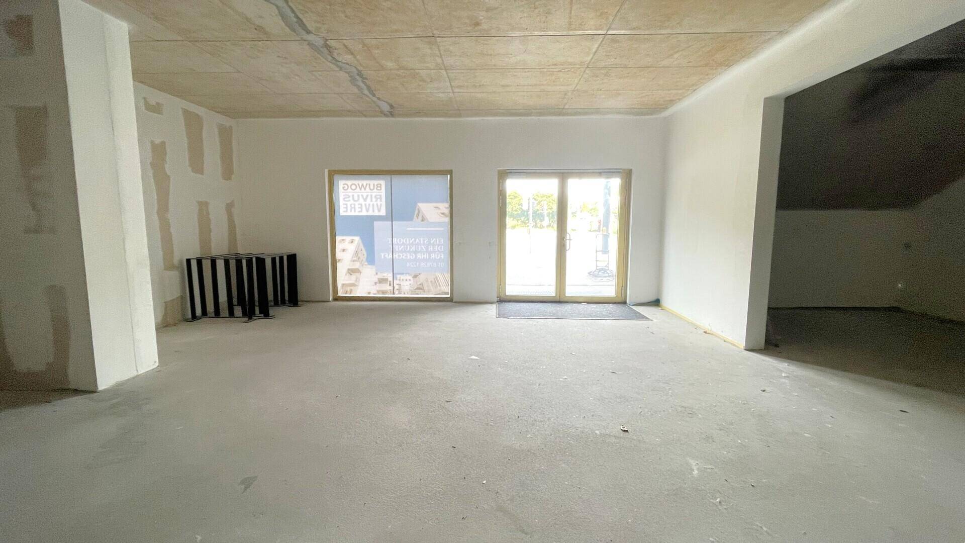 Unfertiger Innenraum mit großen Glastüren und Fenstern, die Tageslicht hereinlassen, und einem Betonboden.