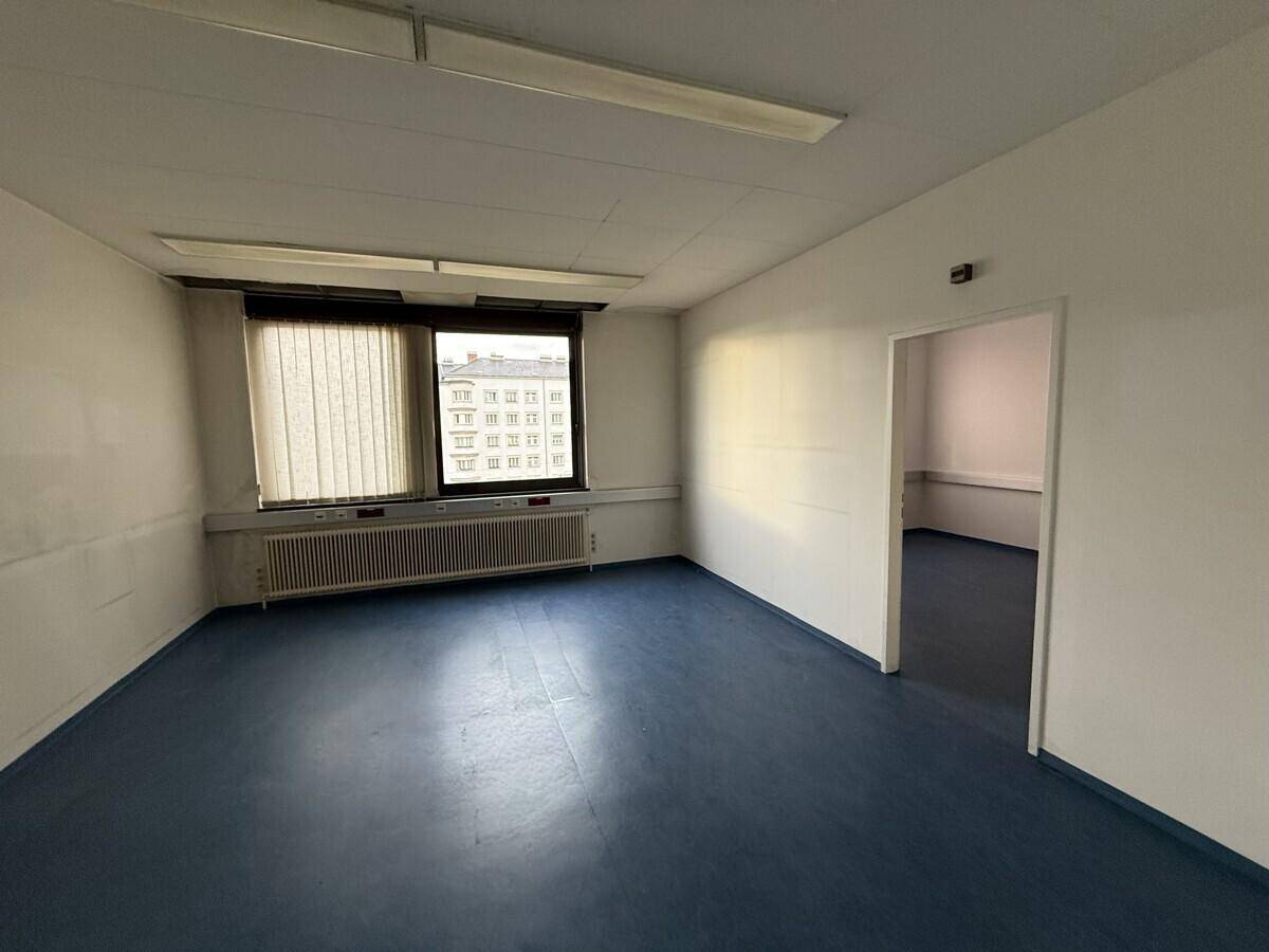 Leerer Büroraum mit blauen Vinylböden, einem großen Fenster und Zugang zu einem weiteren Bereich.