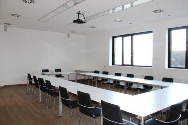 Heller Konferenzraum mit flexibler Bestuhlung, Projektor und großen Fenstern für eine angenehme Atmosphäre.
