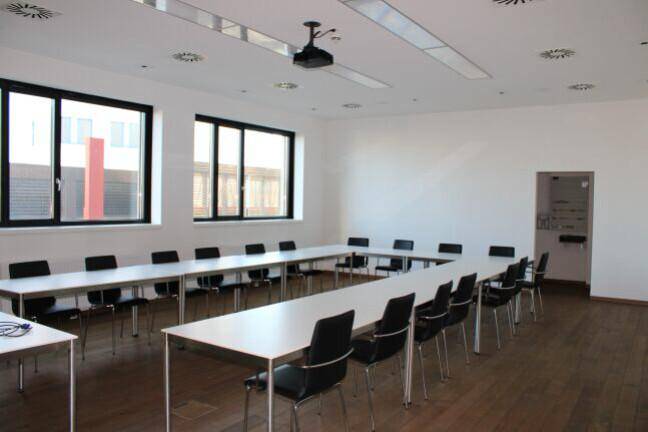 Großer Konferenzraum mit U-förmiger Tischordnung, Projektor und großen Fenstern für Tageslicht.
