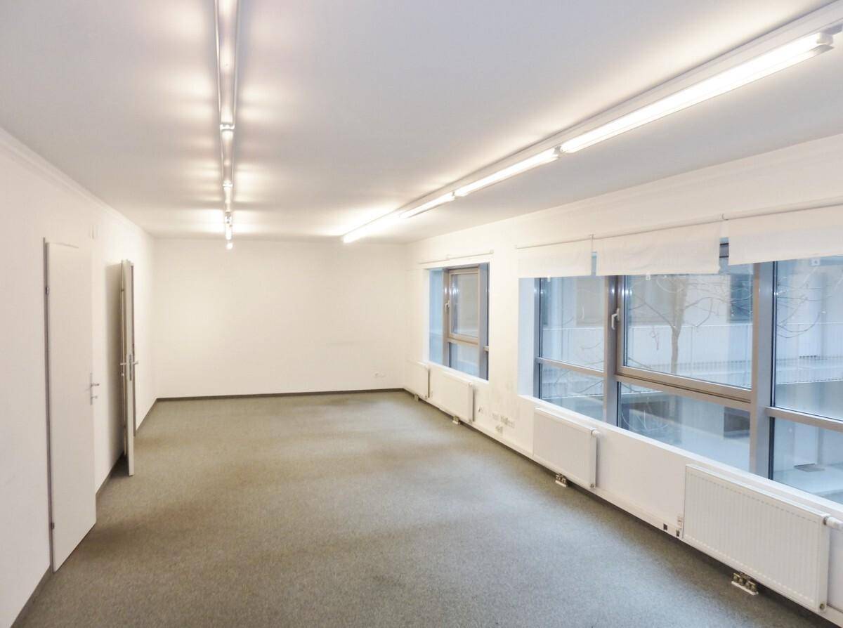 Leerer, heller Raum mit Teppichboden, großen Fenstern und Deckenbeleuchtung, ideal für Büronutzung.