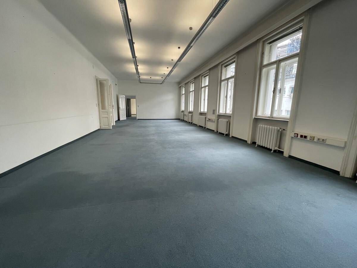Geräumiger Bürobereich mit durchgehender Fensterfront, blauem Teppich und Deckenleuchten, ideal für Großraumbüros.