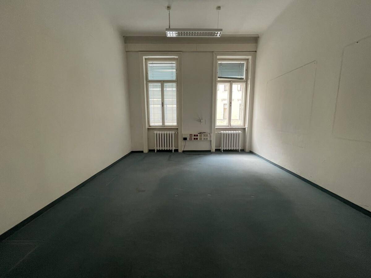 Heller Büroraum mit zwei großen Fenstern, blauem Teppichboden und klassischen Heizkörpern.