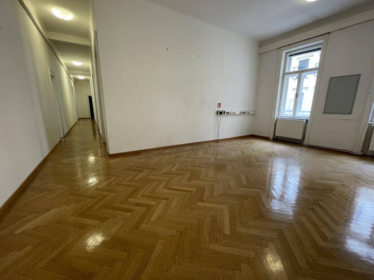 Geräumiger Raum mit glänzendem Fischgrätparkett und Blick in einen langen Flur, ideal für Bürozwecke.