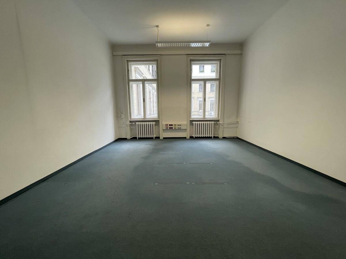 Heller Büroraum mit blauem Teppichboden, zwei Fenstern und einfacher Deckenbeleuchtung, funktional und sauber.