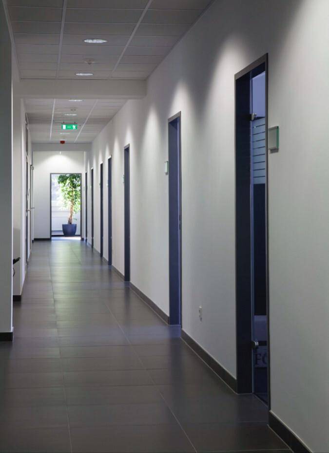 Langer Bürogang mit mehreren Türen auf beiden Seiten, dunklem Fliesenboden und einer Pflanze am Ende.