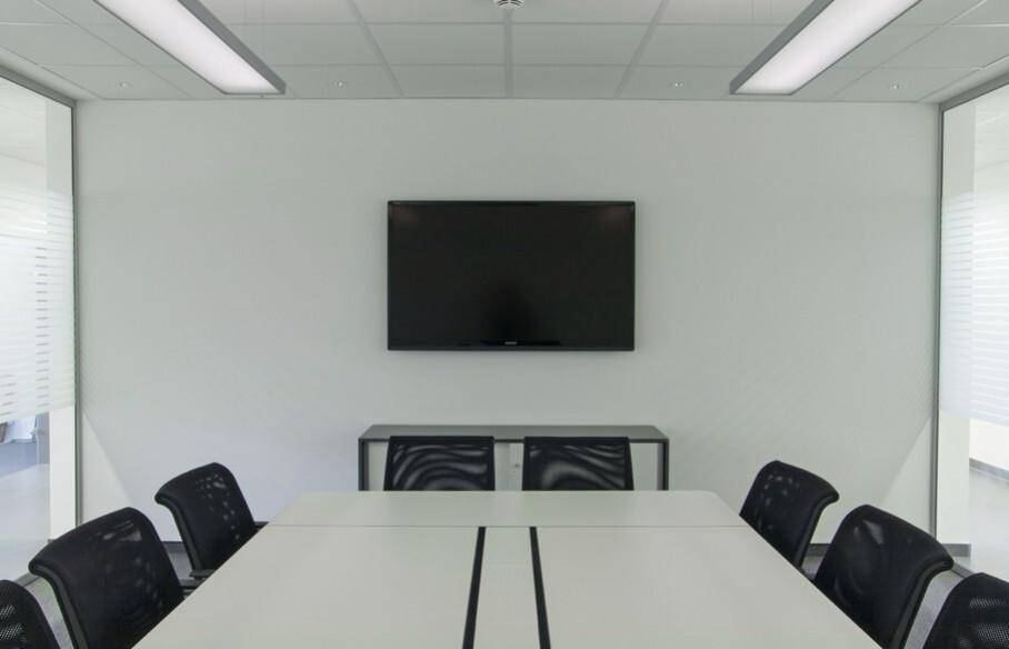Konferenzraum mit langem weißen Tisch, schwarzen Stühlen und einem an der Wand montierten Fernseher unter Leuchtstoffröhren.