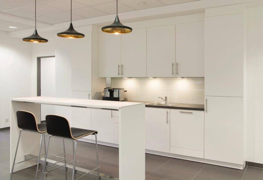 Moderne Teeküche mit weißen Schränken, Spüle und einer Bar mit zwei Hockern, beleuchtet von Pendelleuchten.