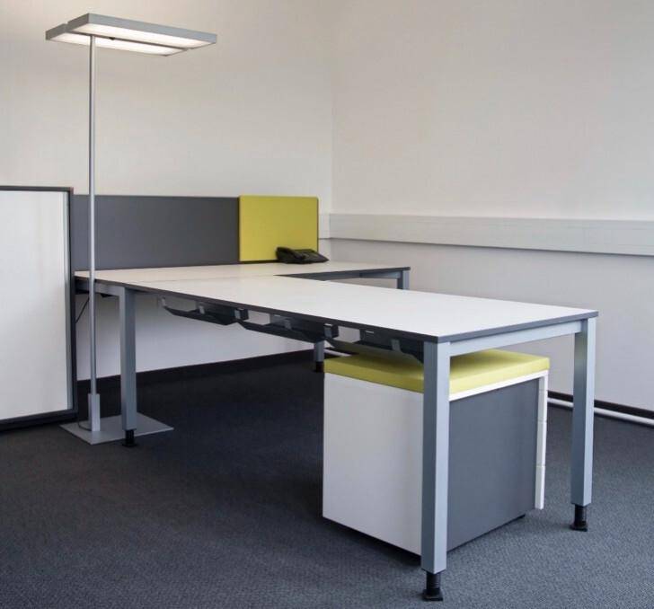 Moderner Büroarbeitsplatz mit L-förmigem Schreibtisch, Rollcontainer und einer Stehlampe auf grauem Teppichboden.