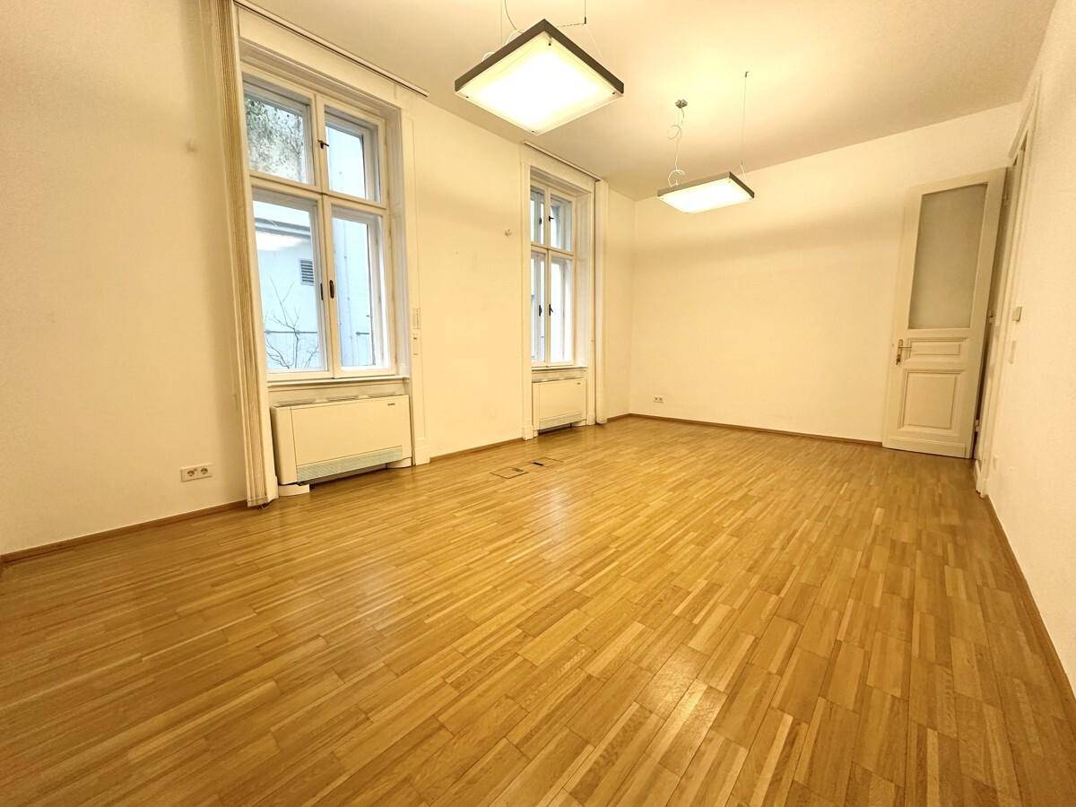 Heller Büroraum mit Holzboden, hohen Fenstern und weiß gestrichenen Wänden.
