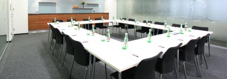 Konferenzraum mit U-förmigem Tisch, schwarzen Stühlen und Teppichboden, ideal für Besprechungen.
